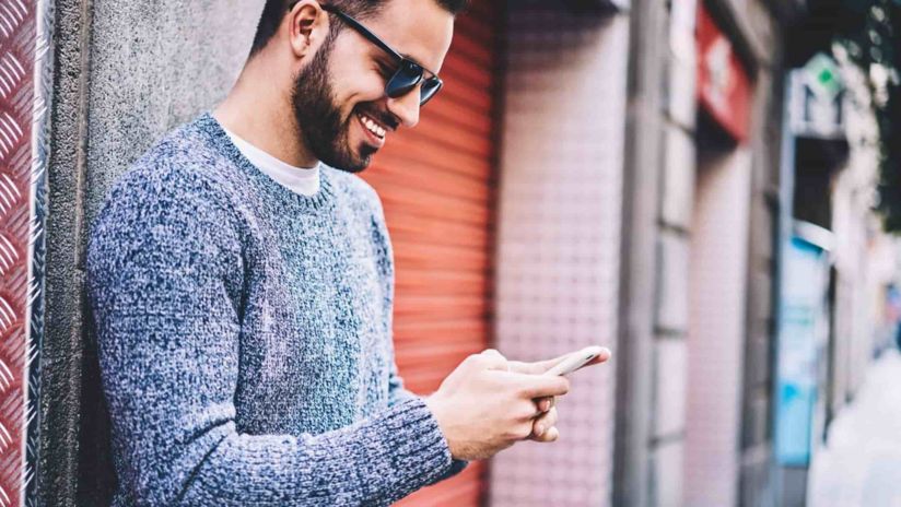 A man wearing sunglasses and a grey jumper smiles as he looks down and types on his smartphone.