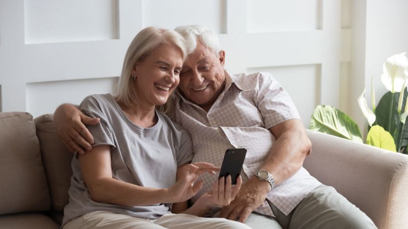 An older couple smile as they sit together on a lounge and look down at something on a smartphone screen.