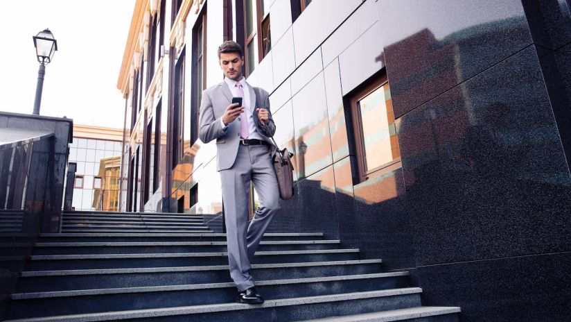 A man wearing a suit looks down at his smartphone as he walks down a set of stairs outside in an urban environment.